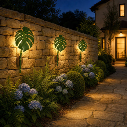 Three Linin Monstera leaf outdoor wall lights lit along a stone garden wall at nightfall | Wickleigh